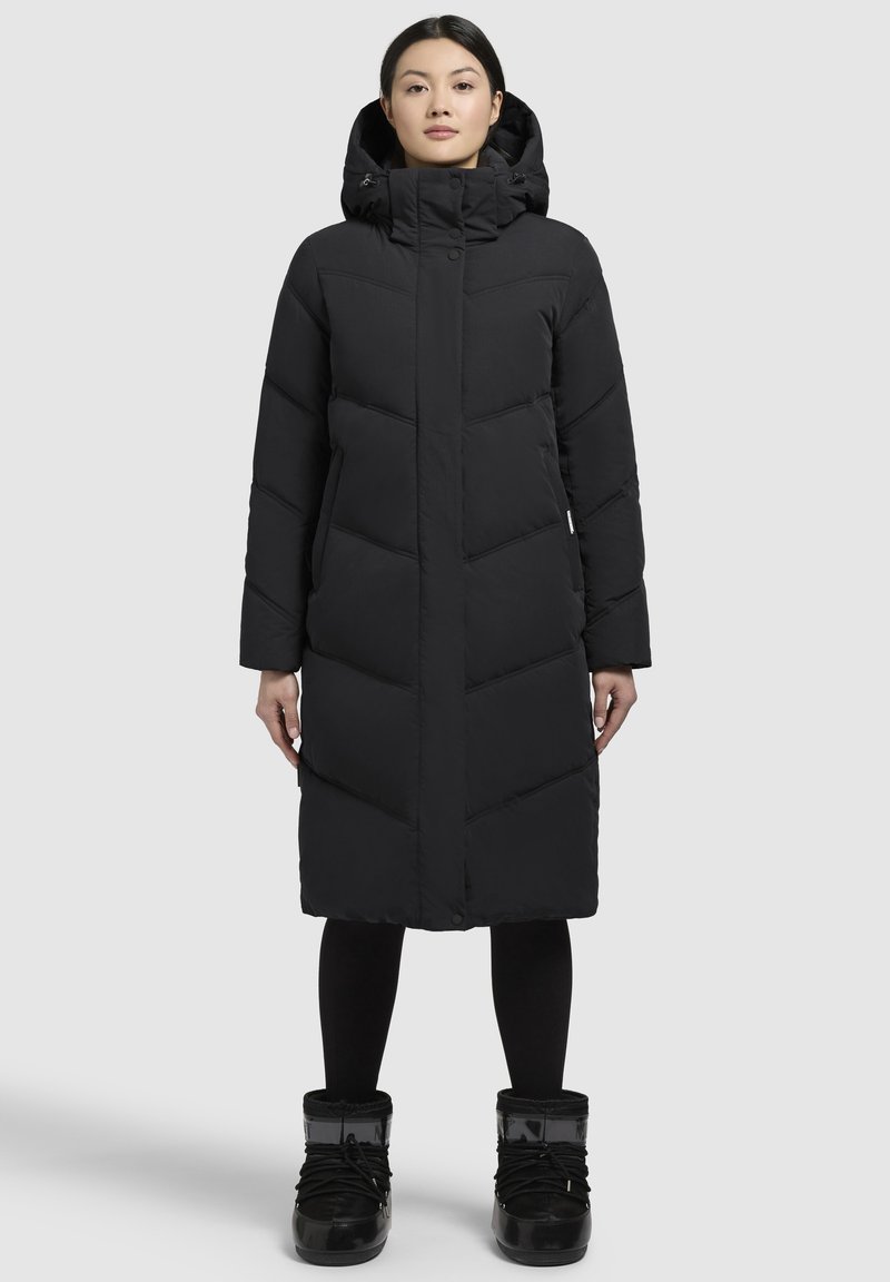 Caban lung negru cu glugă, prezentând cusături în formă de V, închideri cu capse/ butoane și buzunare laterale. Asortat cu cizme negre de zăpadă.