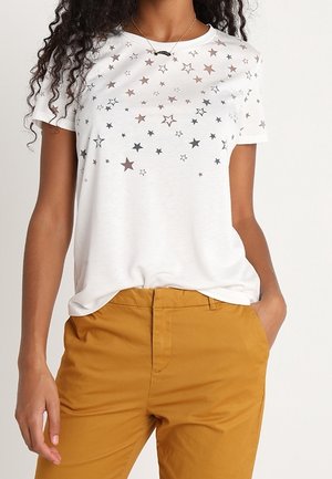 Femme portant un t-shirt blanc à manches courtes avec divers imprimés d'étoiles argentées et un pantalon jaune moutarde, une main dans la poche.
