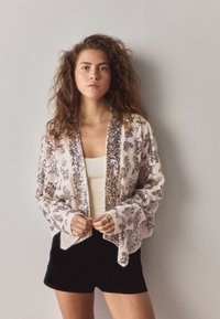 Beige sheer jacket with silver sequins, open front and wide sleeves, worn over a white tank top and black shorts. Soft texture, adorned detailing.