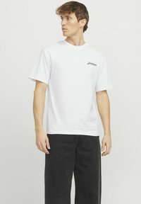Camiseta blanca de algodón con mangas cortas, corte holgado, que presenta un pequeño logotipo negro en el pecho. Combinada con unos vaqueros anchos negros.