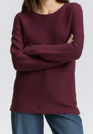 Personne portant un pull côtelé à manches longues de couleur bordeaux foncé, les bras croisés, associé à un jean bleu en denim sur un fond uni.