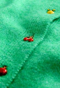 Țesătură tricotată verde cu butoane decorative în formă de cireșe roșii, un buburuză roșie și o albină galbenă.
