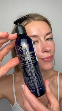 Frau mit heller Haut und blondem Haar hält eine blaue Flasche Kiehl's Midnight Recovery Botanical Cleansing Oil nah an ihr Gesicht.