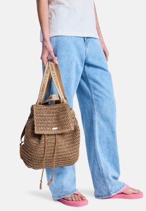 Ruskea punottu reppu, jossa on läppä ja kiristysnauha, logovyö ja teksturoitu pinta. Yhdistetty vaaleansinisiin farkkuihin ja vaaleanpunaisiin sandaaleihin.