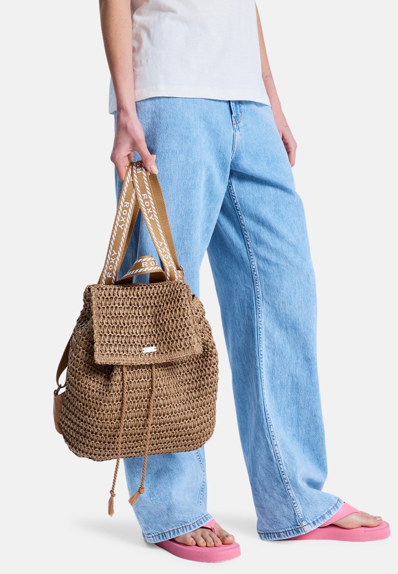 Καφέ υφαντό σακίδιο με καπάκι και κλείσιμο με κορδόνι, με φανερή ράβδο με λογότυπο και υφή στην επιφάνεια. Συνδυασμένο με ανοιχτόχρωμο τζιν και ροζ σανδάλια.