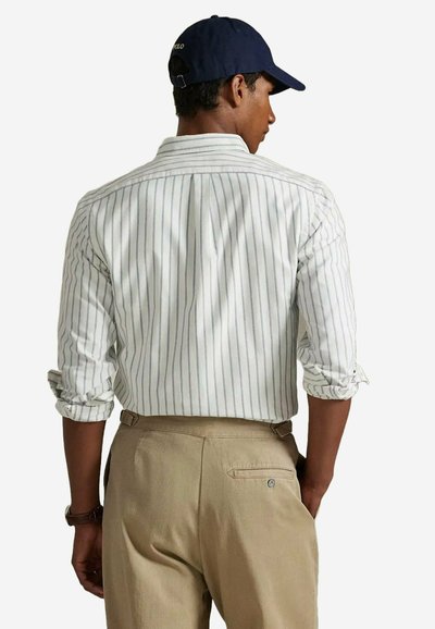 Chemise à rayures claires en blanc et gris, avec des manches retroussées. Pantalon beige avec un détail de poche arrière. Casquette navy complète le look.