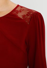 Červené tričko s dlouhým rukávem, které se vyznačuje texturovanou tkaninou, krajkovým překryvem na ramenou, plisovanými detaily a kulatým výstřihem se hladkým povrchem.