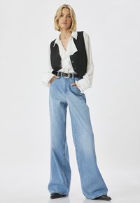 Chaleco negro sin mangas sobre una camisa blanca con volantes, combinado con jeans de pierna ancha de color azul claro y un cinturón negro con hebilla de plata. Texturas suaves.