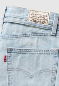 Lyseblå denimjeans med sydd baklomme og en rød Levi's-lapp. Inkluderer et stoffmerke med merkenavn og grafisk design.