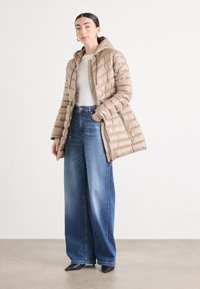 Giacca a vento trapuntata beige con cappuccio, con cuciture orizzontali e tasche con zip, indossata sopra un top bianco, abbinata a jeans blue a gamba larga.