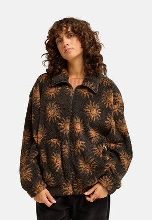 Femme portant une veste zippée noire et marron duveteuse avec des motifs de rayons de soleil, debout devant un fond clair uni.