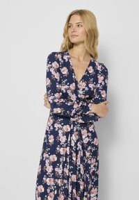 Sukienka w kwiaty z falbaną w granatowym materiale, ma długie rękawy, głęboki dekolt w kształcie litery V oraz marszczoną talię w różowe i białe wzory kwiatowe.