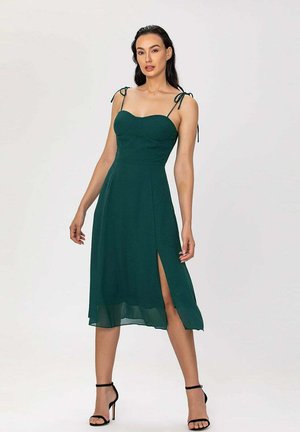 Femme posant dans une robe midi sans manches vert foncé avec une fente latérale et des sandales noires à talons hauts avec bride à la cheville sur fond uni.