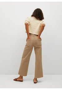 Femme aux cheveux bouclés portant un pull en tricot crème, un pantalon large beige avec poches arrière et des sandales à bout ouvert marron, se tenant contre un mur blanc.