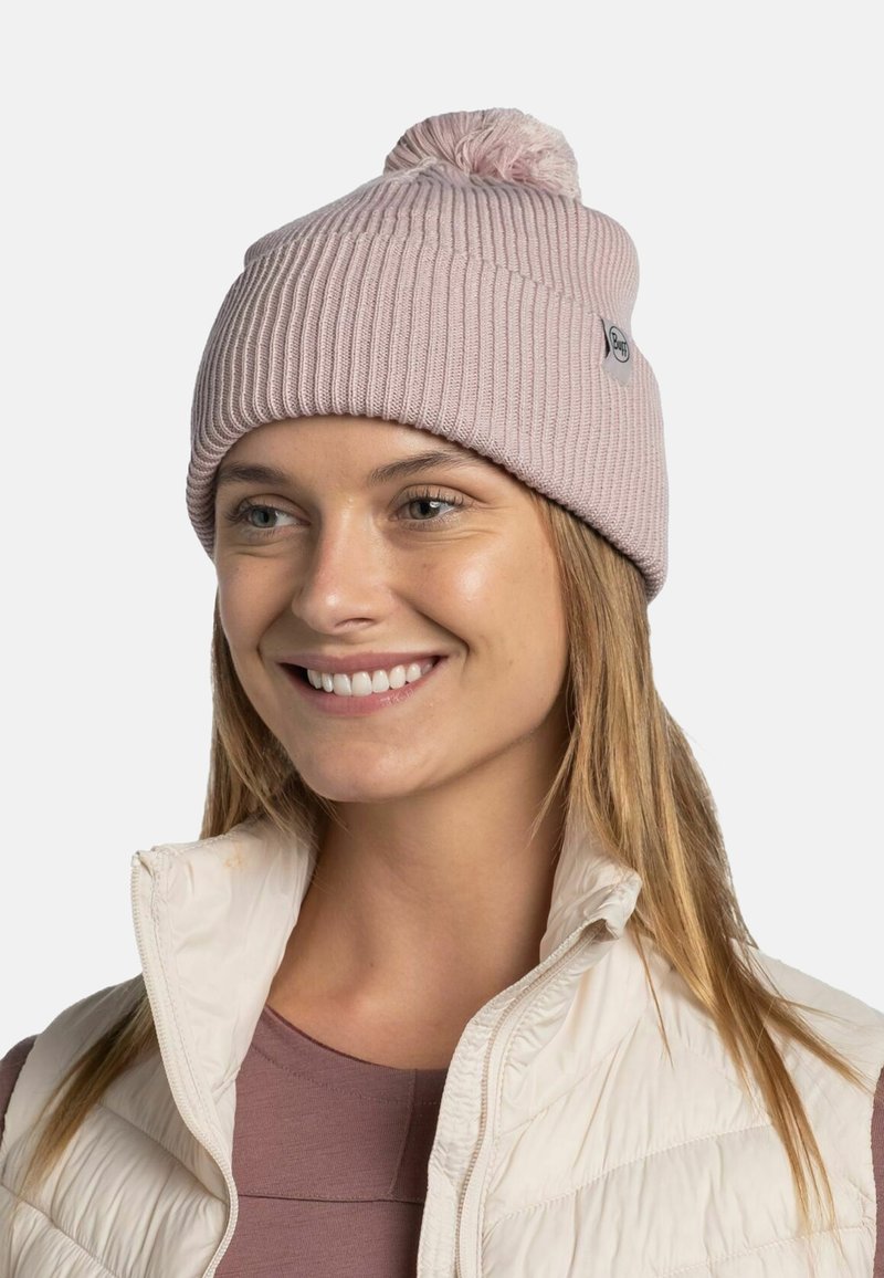 Light pink ribbed knit beanie with a pom-pom on top, features a small logo tag, worn with a white puffy vest and a mauve top underneath.