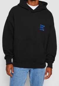 Sudadera con capucha negra hecha de algodón suave con un bolsillo tipo canguro. Cuenta con un logo azul de "Calvin Klein Jeans" en el lado izquierdo. Textura de caucho.