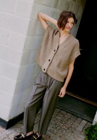 Gilet lavorato a maglia beige con scollo a V, maniche corte e bottoni scuri, abbinato a pantaloni grigi su misura e scarpe marroni. Superfici testurizzate visibili.