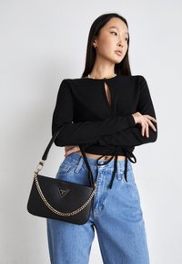 Czarna torba crossbody z łańcuszkiem, teksturowane wykończenie, trójkątne logo, zestawiona z czarną bluzką i jasnoniebieskimi dżinsami.