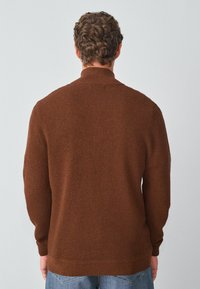 Brązowy sweter robiony na drutach z teksturowanym wzorem, wysokim kołnierzem oraz ściągaczami na mankietach i dole, widziany z tyłu. Dopełniony niebieskimi dżinsami.