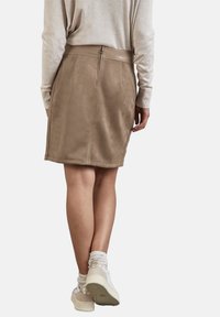 Gonna in finta suede marrone con un design aderente, di lunghezza media, e chiusura con zip sul retro. Abbinata a una maglietta beige e sneakers chiare.