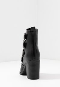 Botín negro de tacón alto hecho de cuero sintético, con hebillas plateadas y un tacón texturizado. Superficie suave con punta redonda.