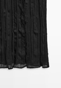 Czarna przezroczysta spódnica z pionowymi, fakturowanymi paskami i lekko marszczonym dołem na białym tle.