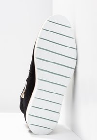 Chaussure noire avec une semelle blanche épaisse présentant de larges rainures horizontales, placée semelle vers le haut contre un mur blanc d’angle.