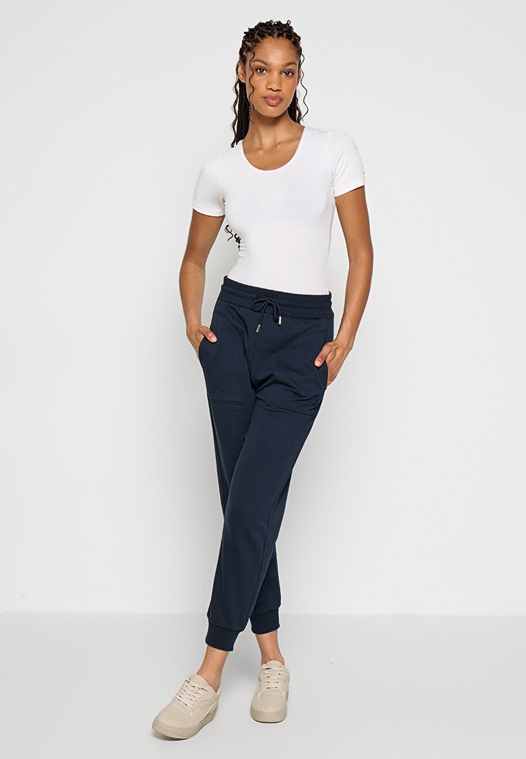 Weißes, enges T-Shirt kombiniert mit marineblauen Sweatpants, die über zwei Seitentaschen und elastische Bündchen verfügen, getragen mit beigen Turnschuhen. Glatte Stoffoberfläche.