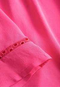Blusa rosa con un acabado texturizado, que presenta un galón de encaje decorativo en el dobladillo de las mangas. La tela es ligera y suave.