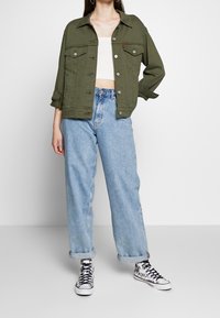 Veste en denim vert olive avec fermeture à boutons, deux poches poitrine, associée à un jean taille haute bleu clair à jambes larges et des baskets noires et blanches à motifs.