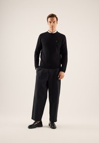 Pull noir avec des poignets côtelés, associé à un pantalon noir ample et des chaussures en cuir noires. Détail de col blanc simple visible.