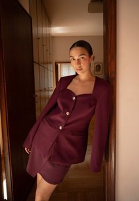 Burgundy räätälöity bleiseri pitkillä hihoilla, neliönmuotoinen pääntie, yksittäinen nappisulku ja siihen sopivat hame; sileä tekstuuri ja jämäkkä muotoilu.