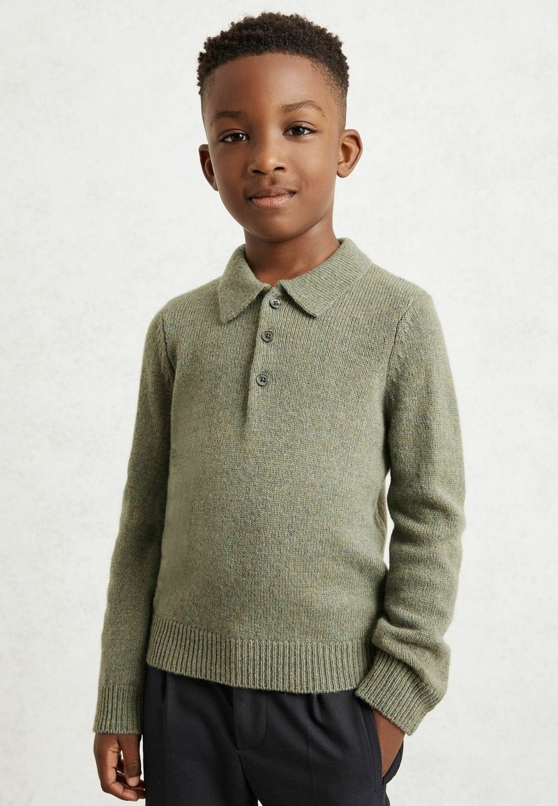 Zöld kötött pulóver galléros nyakkal, gombos záródással és bordázott mandzsettákkal. Sötét nadrággal viselve, világos háttér előtt.