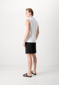 Camisa polo sin mangas blanca, pantalones cortos negros y sandalias negras. El conjunto presenta un diseño simple y una textura suave.