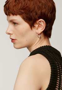 Prenda de punto negra con diseño texturizado; cabello corto y vibrante de color rojo; pendiente de aro de plata; vista de perfil lateral.