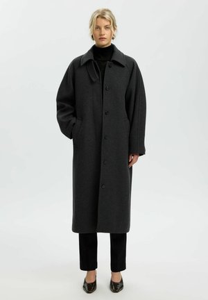 Un mannequin blond se tient devant un fond de studio clair, vêtu d’un long manteau boutonné gris anthracite par-dessus des vêtements et des chaussures noirs.