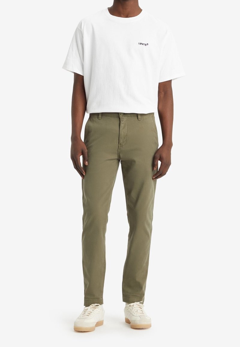 Camiseta blanca de algodón con un pequeño logo negro de Levi's, combinada con pantalones chinos verde oliva y zapatillas blancas con suela beige. Diseño sencillo.