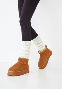 Bottines en daim marron avec des semelles en caoutchouc, portées sur des jambières en laine blanches, associées à des leggings noirs.