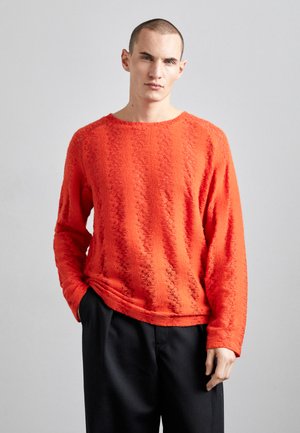 Pulover roșu texturat, cu guler rotund și mâneci lungi, asortat cu pantaloni negri. Tesătura are un model unduit și o croială confortabilă.