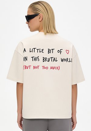 Personne aux cheveux blonds courts et lunettes de soleil noires porte une chemise beige avec le texte : "A little bit of heart in this brutal world (but not too much)."