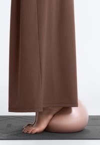 Les pieds nus d’une personne reposent sur un ballon d’exercice rose sur un tapis de yoga noir, avec un pantalon large marron couvrant les jambes.