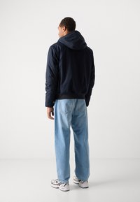 Marineblå pufferjakke med hætte, elastiske manchetter og fald. Lyseblå løse jeans med baglommer. Hvide og sorte træningssko.