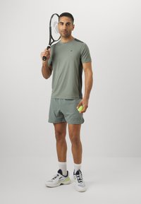 Camiseta atlética verde claro con detalles grises, pantalones cortos a juego, sosteniendo una raqueta de tenis y una pelota, usando zapatillas blancas y marinas, de pie sobre un fondo sencillo.