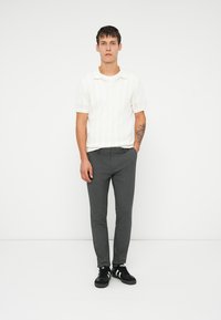 Mladý muž s tetováním na levém předloktí má na sobě bílé tričko s krátkým rukávem, šedé slim-fit kalhoty a černé tenisky s bílými pruhy.