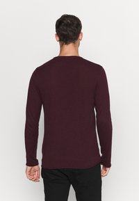 Man som vänder bort med ryggen, iförd en enfärgad långärmad mörk vinröd skjorta och svarta byxor mot en vit bakgrund.