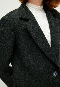 Blazer di lana nera con texture, dotato di colletto a lancia e caratterizzato da un singolo bottone blu, indossato sopra un top bianco a coste.