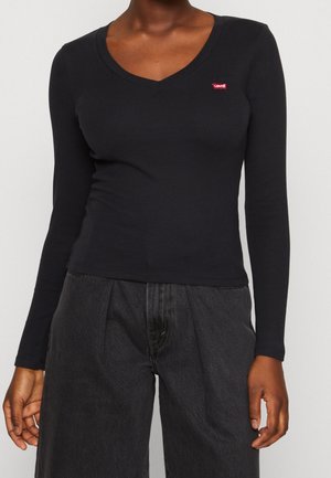 Femme portant un haut noir cintré à manches longues et col en V avec un petit logo rouge Levi's sur la poitrine et un pantalon gris foncé ample.