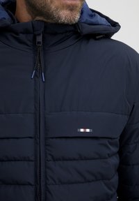 Jachetă de tip puffer de culoare bleumarin, cu fermoar pe față, dotată cu glugă reglabilă, textură matlasată și un mic logo cu accente roșii, albe și gri.