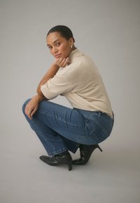 Beež lühikeste varrukatega särk, sinised teksad ja mustad terava ninaga kontsakingad. Modell kükakil, üks käsi lõual, neutraalne taust.