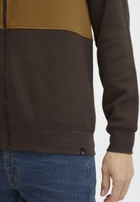 Braune und senfgelbe Zip-Jacke mit kontrastierenden horizontalen Paneelen, weicher Textur, gerippten Bündchen und einem kleinen Markenetikett am unteren Ende.